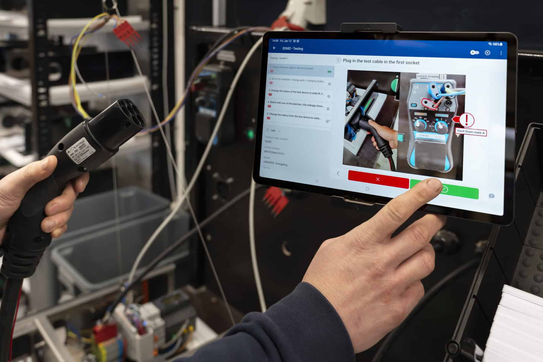A person using a tablet for instructions while holding a cable plug. The screen displays assembly steps for connecting the plug to a socket, including images and text. The setting appears to be a technical or workshop environment with various equipment in the background.