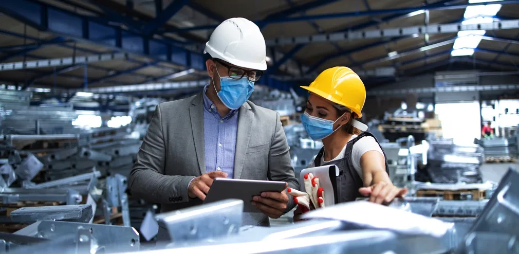 Two workers wearing safety helmets and face masks examine documents and a tablet in a large industrial warehouse. The man on the left wears a white helmet and suit, while the woman on the right wears a yellow helmet and safety vest, surrounded by metal equipment.