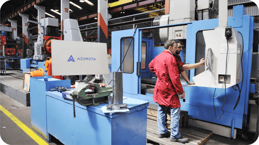 Zwei Arbeiter bedienen eine große Maschine in einer Industriehalle. Der eine in einem roten Mantel schaut auf den Bildschirm, während der andere die Bedienelemente einstellt. In der Nähe sind verschiedene Maschinen und Werkzeuge, darunter ein Schraubstock, zu sehen. Eine 