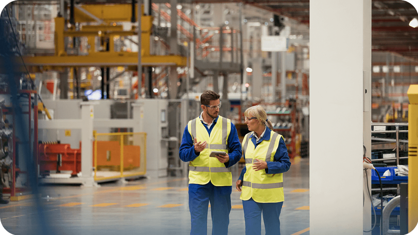 Two workers in high-visibility vests walk through a large industrial facility, engaging in conversation. The facility is well-lit and contains various machinery and equipment. Both appear to be focused, with one holding a tablet.