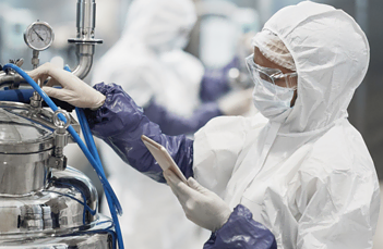 A person wearing a protective suit, gloves, mask, and hairnet operates a piece of industrial equipment, possibly in a laboratory or manufacturing facility. They are reading a document and turning a valve on a metallic container amidst a blurred background.