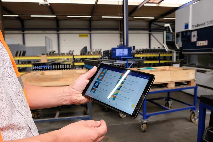 A person holds a tablet displaying colorful designs and data while standing in an industrial workshop. The background includes machinery, including a large press brake labeled 