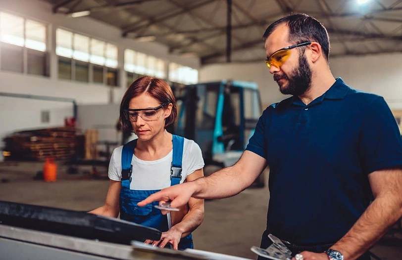 Two factory workers wearing safety goggles, one male and one female, collaborate in an industrial setting. The female worker, wearing overalls, operates a machine with a touchscreen while the male worker points at the screen, both focused on the task.