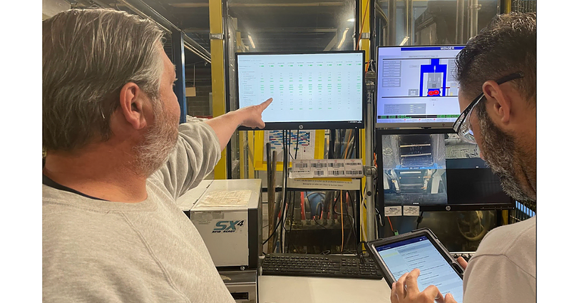 Twee mannen bekijken informatie op verschillende schermen in een controlekamer. De ene man wijst naar een groot scherm met gedetailleerde gegevens, terwijl de andere een tablet vasthoudt. De kamer heeft industriële apparatuur en verschillende technische displays.