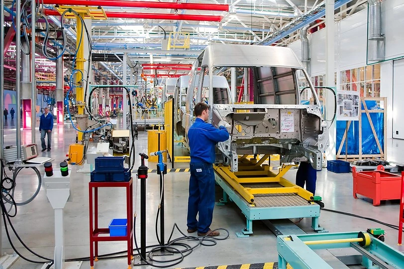 Workers in blue overalls are assembling a large vehicle in a bright, organized factory setting. Mechanical parts and equipment surround the production line, with colorful piping overhead and various workstations along the assembly path.