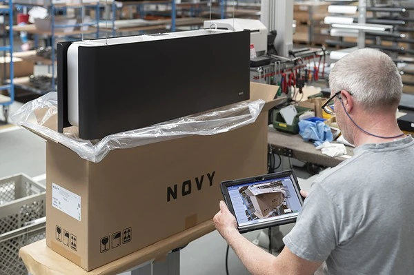 A person with gray hair and glasses stands in a factory setting, holding a tablet. In front of them, there's a large box labeled 