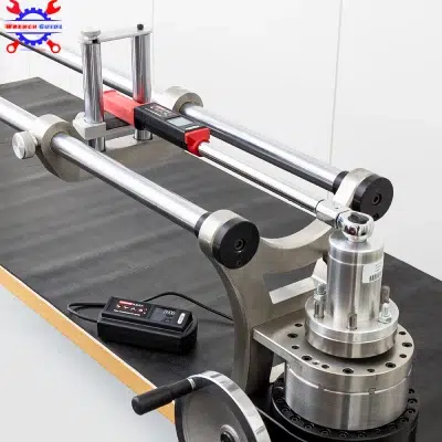 A precision torque wrench calibration setup is shown. It consists of dual horizontal metal rods supporting a central mechanism with a digital gauge, mounted on a sturdy base. An electronic control unit is placed to the side. The workspace surface is black.