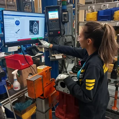 A female factory worker with a ponytail operates a machine while referencing information on a large monitor. She is wearing safety glasses and gloves. The workspace is equipped with various tools, parts, and machines.