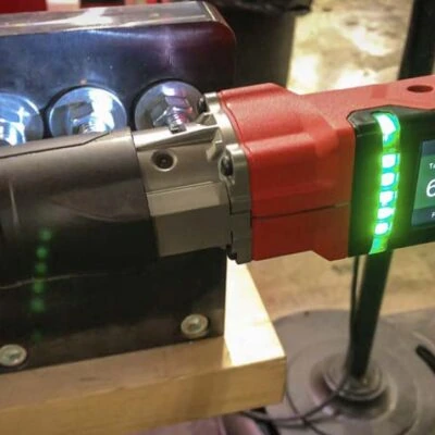 A battery-powered electric ratchet tool with a digital display and LED light indicators is set up on a workbench. The tool has a red and black casing and is secured in place. Adjacent to it are a few metallic fixtures and a blurred background.
