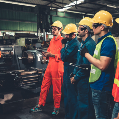 A group of factory workers in protective gear, such as yellow hard hats and safety glasses, listen attentively to an instructor in a workshop. They stand near industrial machinery, with some workers holding tablets and notebooks.