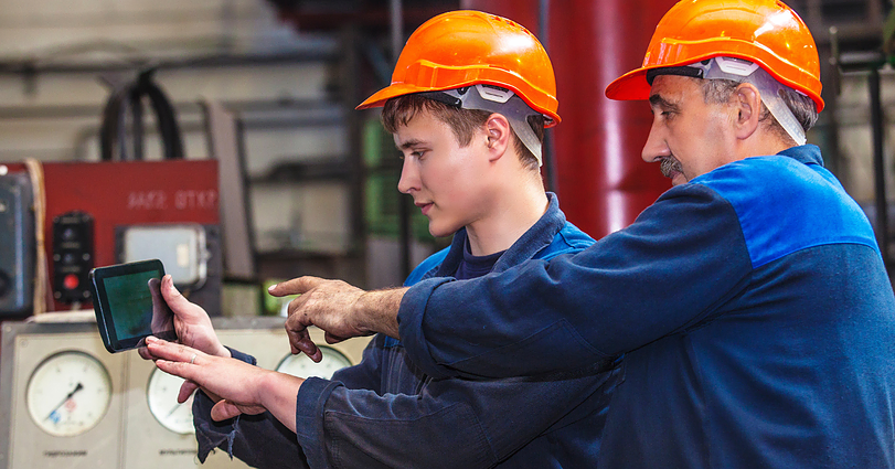 Two workers in blue overalls and orange helmets are using a tablet in an industrial setting. The older worker points at the screen, guiding the younger worker. Gauges and industrial equipment are visible in the background.