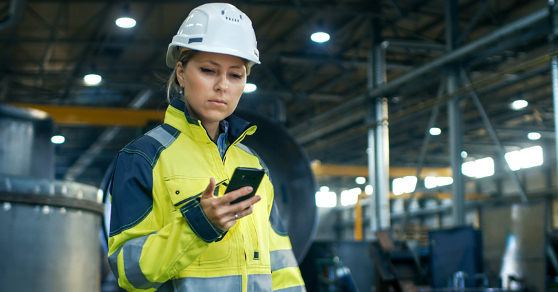 A person in a yellow and gray high-visibility jacket and a white hard hat is looking at a smartphone. They are standing inside what appears to be an industrial facility with machinery and metal structures in the background.