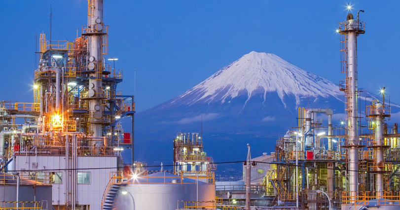 An industrial factory with complex machinery and towers stands in the foreground, lit with bright lights. In the background, Mount Fuji with its snow-capped peak is visible against a blue sky, creating a striking contrast between nature and industry.