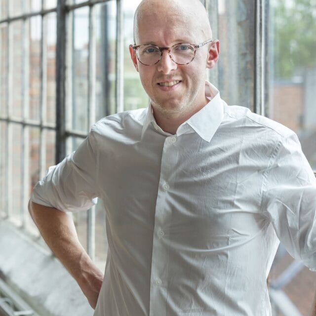 A person with a shaved head and glasses, wearing a white button-up shirt, stands by large industrial-style windows with one hand on their hip, smiling at the camera. The sunlight coming through the windows highlights the scene.