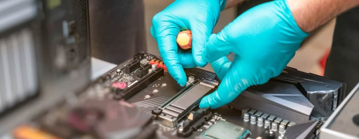 A pair of hands wearing blue gloves is installing or repairing components on a computer motherboard using a screwdriver. The focus is on the hands and the motherboard, with various electronic parts visible.