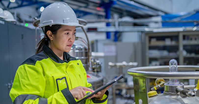 A woman in a safety helmet and high-visibility jacket is working in an industrial environment. She is holding a tablet and inspecting equipment or machinery in a factory setting. Various industrial components and machinery are visible in the background.