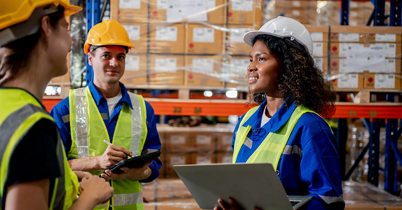 Three warehouse workers wearing safety vests and hard hats are having a discussion. One woman is holding a laptop, while another woman and a man are taking notes on clipboards. Shelves filled with boxes are visible in the background.