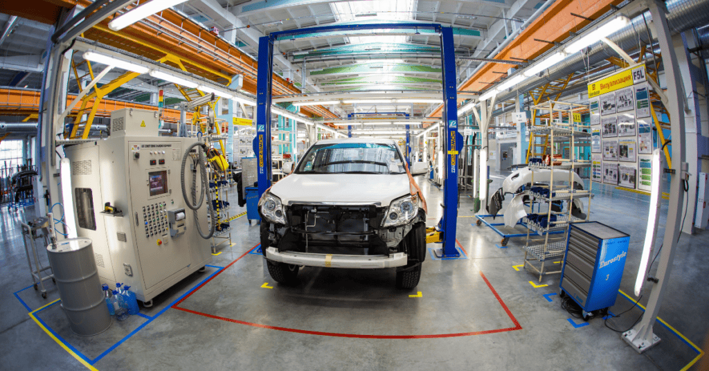 A car assembly line in a factory with machinery and equipment. A partly assembled car is centered in the frame, with components and tools organized around it. The setting is well-lit, and the industrial space appears clean and orderly.