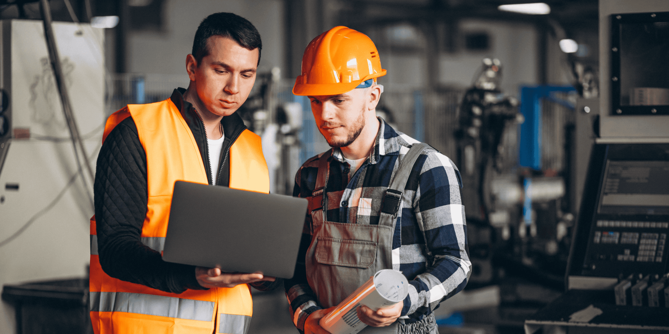 Two men in a factory setting. One is wearing an orange safety vest over black clothes and holding a laptop, while the other is wearing a hard hat and checkered shirt, holding papers. They are engaged in a discussion, possibly reviewing plans or data on the laptop.