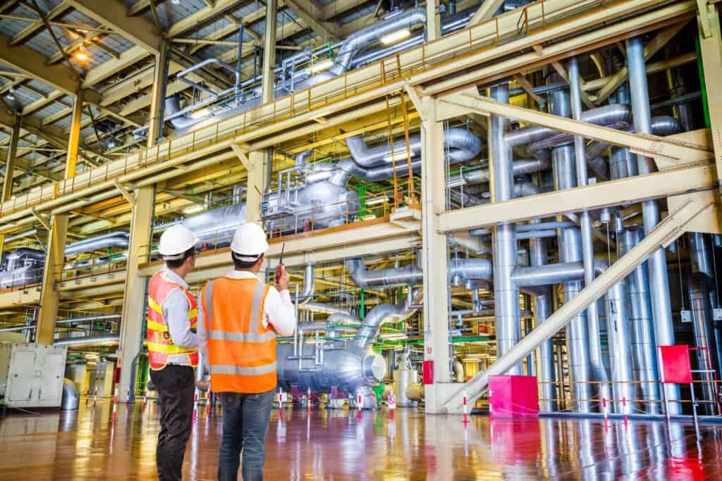 Two workers in orange safety vests and white helmets stand inside a large industrial facility. They are observing and discussing the complex network of pipes and machinery that fill the spacious, well-lit area.