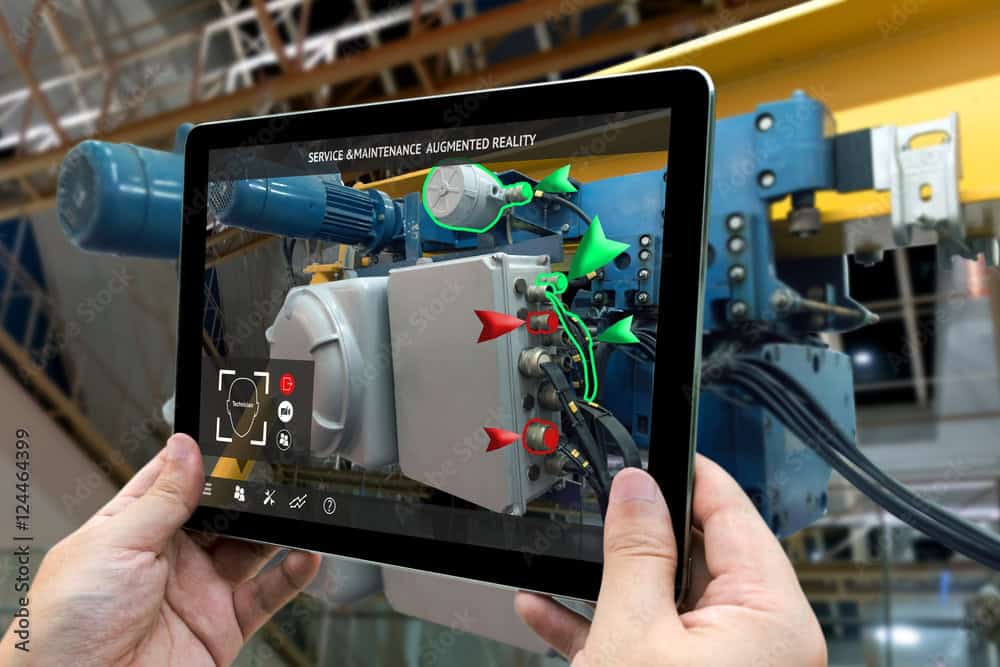 A person holds a tablet displaying augmented reality to perform industrial equipment maintenance. The screen overlays 3D arrows and icons onto machinery, providing guidance and diagnostics, enhancing the service and maintenance process in a factory setting.