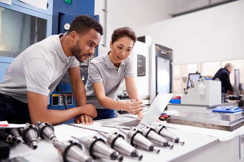 Two engineers in a manufacturing facility, wearing gray polo shirts, working together at a laptop. Various machine components are laid out on the table in front of them. Industrial machinery and another worker are visible in the background.