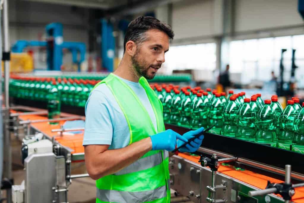 A man wearing a reflective vest and blue gloves holds a smartphone while standing next to a production line filled with green plastic bottles topped with red caps. The industrial setting includes machinery and blurred background elements.