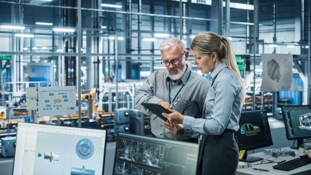 Two engineers, a man with glasses and a woman with a ponytail, review data on a tablet in a high-tech manufacturing facility. Multiple computer screens display 3D models and technical diagrams. The background shows industrial equipment and machinery.