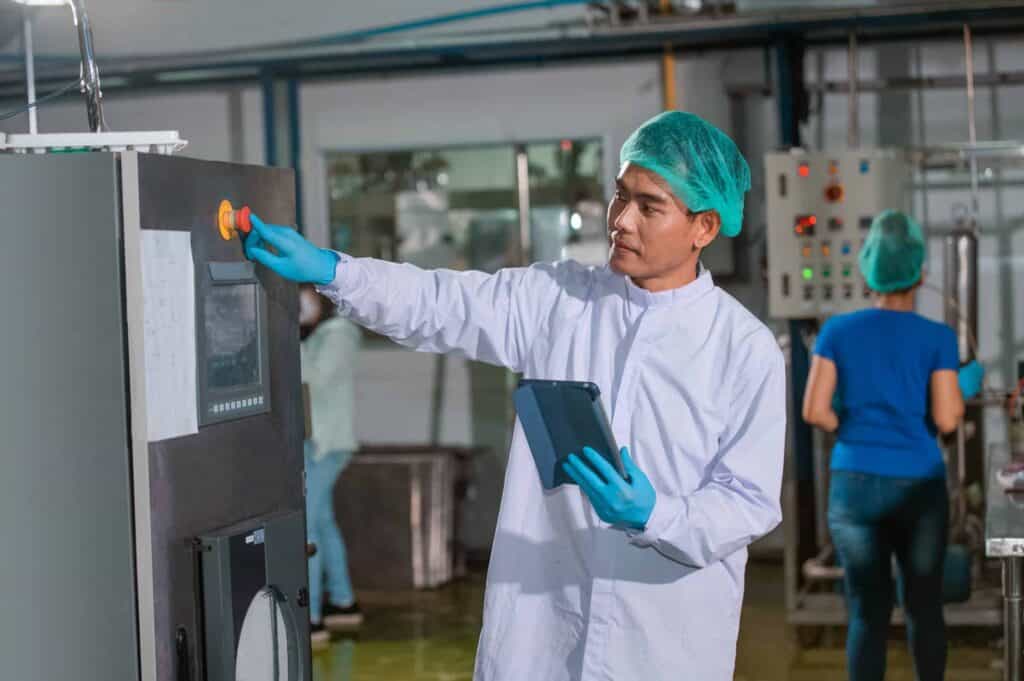 A technician in a white coat and teal hairnet operates machinery with an orange button in an industrial setting. He holds a tablet and wears blue gloves. In the background, another worker with a teal hairnet and blue shirt works at a control panel.