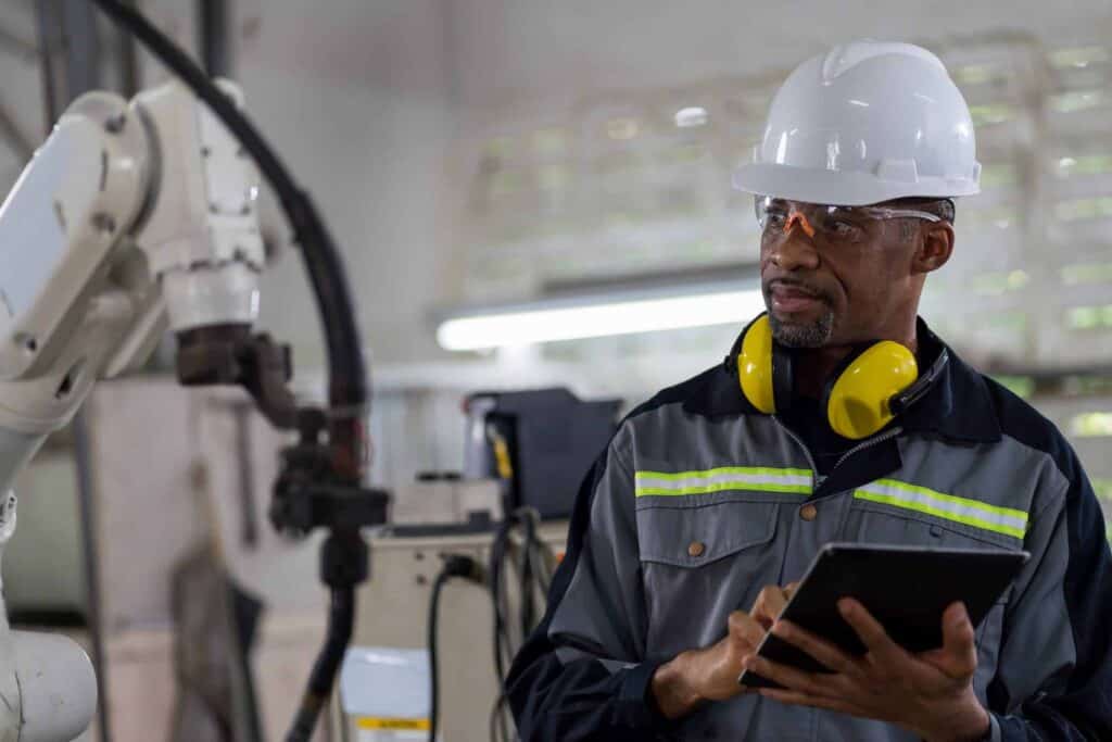 A factory worker in a safety helmet, goggles, and ear protection holds a tablet while inspecting a robotic arm in an industrial setting. The worker wears a reflective safety jacket, and the background shows equipment and machinery.