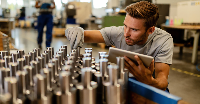 A young man in a workshop uses a tablet while meticulously inspecting rows of cylindrical metal components. He is wearing a grey shirt and a glove on one hand, indicating precision and quality control in a manufacturing environment. Another worker is visible in the background.