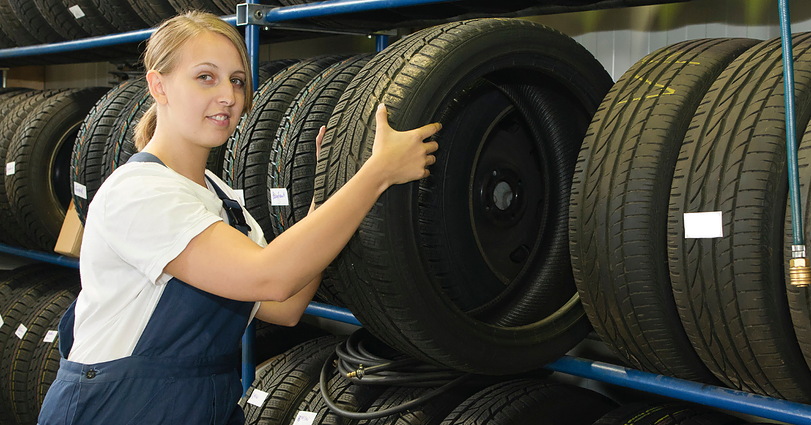 A woman in work overalls arranges a tire on a rack filled with various types of tires in a storage or workshop setting. She is looking at the camera, and the background shows multiple rows of neatly organized tires.