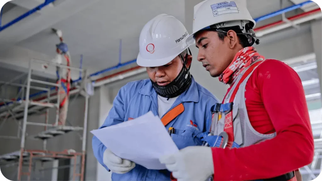Two construction workers wearing safety gear, including hard hats and gloves, are examining a document on a building site. Behind them, a third worker is standing on scaffolding, working on the interior walls. Pipes and construction materials are visible in the background.
