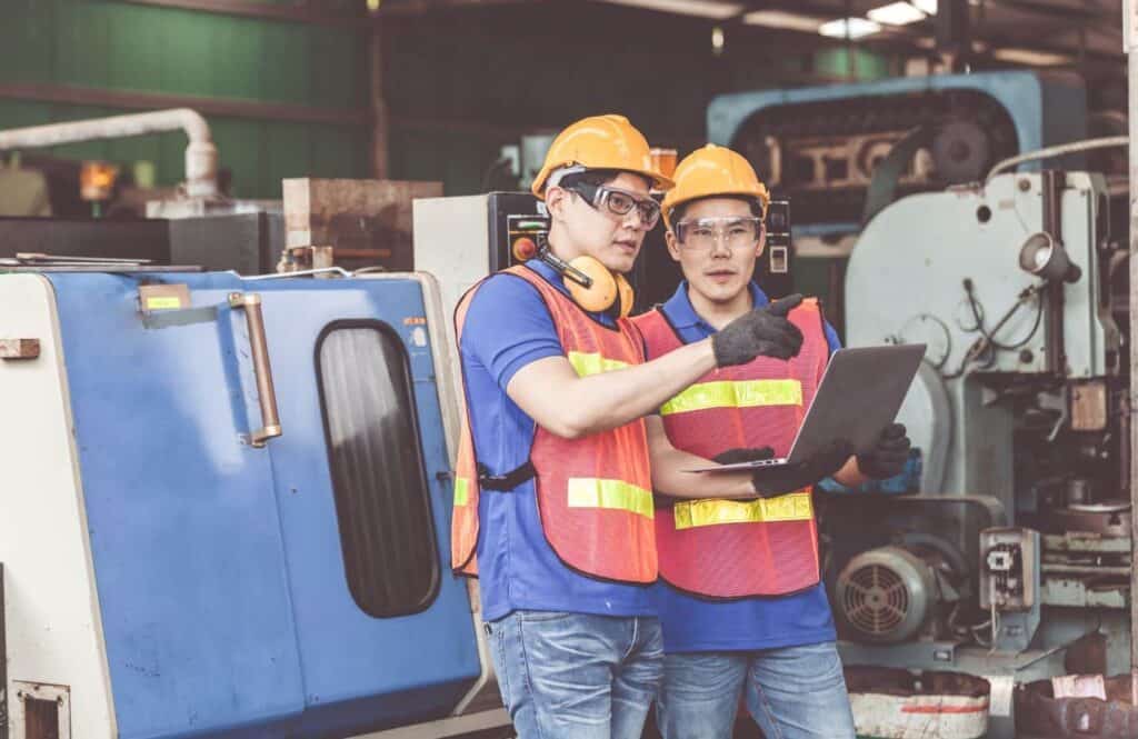 Two industrial workers wearing hard hats, safety vests, and safety glasses, stand together in a factory. One worker is pointing while the other holds a laptop, both appear to be discussing something related to the machinery around them.