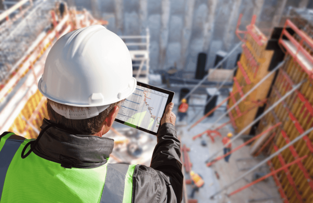 A construction worker wearing a white hard hat and neon green safety vest holds a tablet, while overseeing a construction site with scaffolding and equipment. The tablet screen shows a project plan or schedule. The background is blurred and focused on the worker.