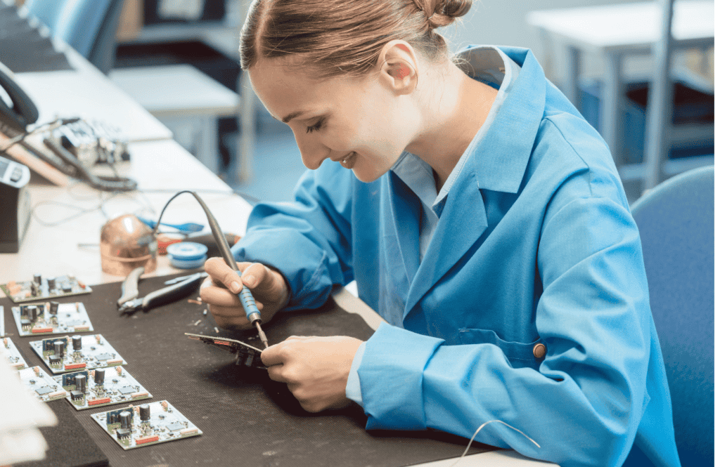 A person wearing a blue lab coat, sits at a worktable soldering electronic components. Various circuit boards and tools are scattered on the table. The individual is concentrated, handling a soldering iron carefully. The background shows more desks and equipment.