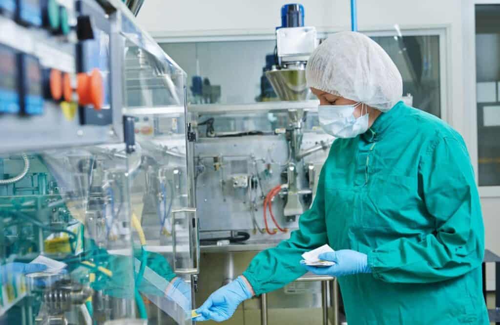 A person in a lab coat, hairnet, and protective mask operates machinery in a sterile laboratory environment. The individual is wearing green protective clothing and appears to be focused on adjusting or monitoring equipment.