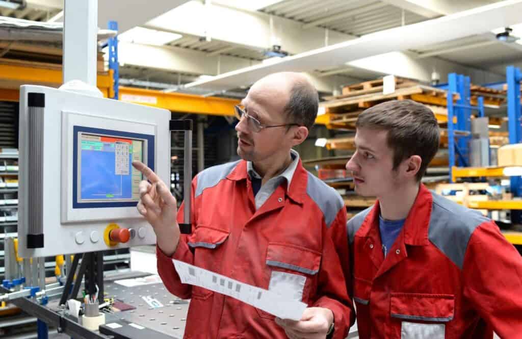 Two factory workers in red and gray uniforms stand in front of a control panel screen. The older worker points at the screen while explaining something to the younger worker, who attentively looks at the display. Both are inside an industrial facility.