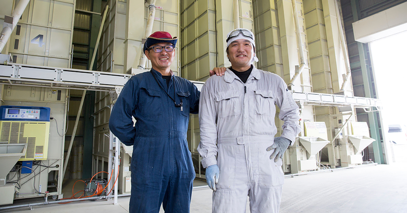 Two men stand side by side, smiling, in front of industrial equipment. One wears a blue jumpsuit, glasses, and a blue and red cap; the other wears a white jumpsuit and a white hard hat. The setting appears to be an industrial or manufacturing facility.