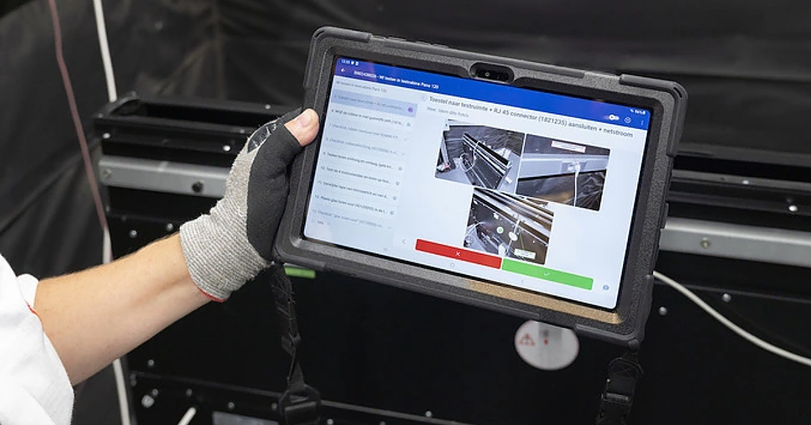 A person wearing a glove holds a tablet displaying architectural designs and annotations. The image on the tablet shows technical diagrams and photos with red, green, and gray annotation indicators. The background includes part of a black and white machine.