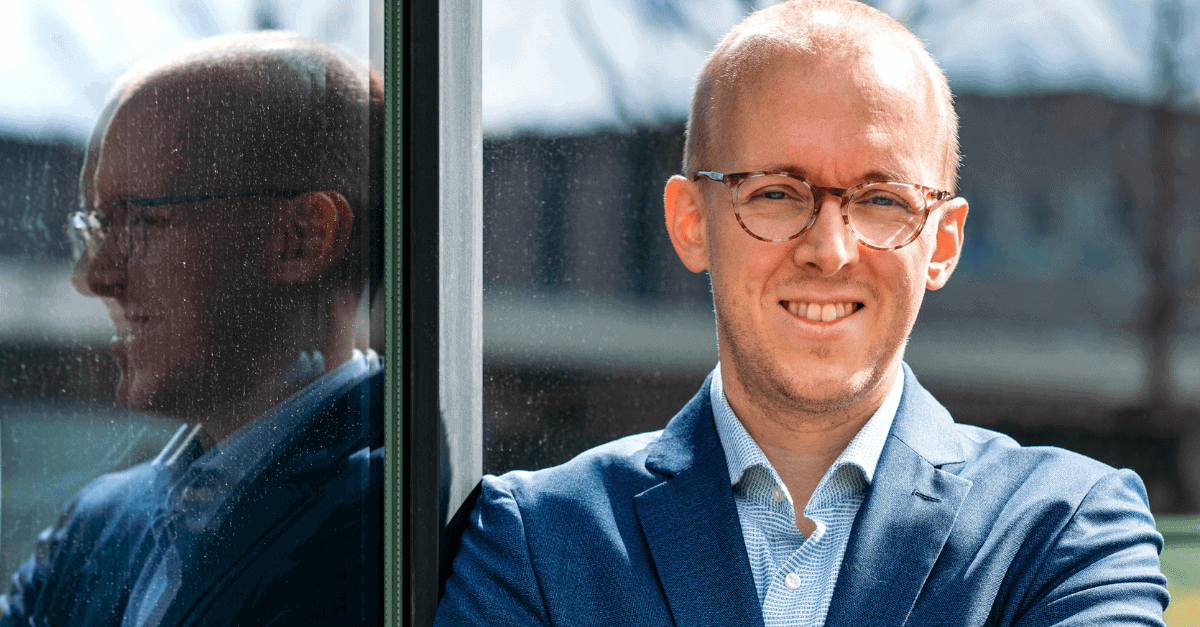 A man with short hair, glasses, and a blue suit smiles while standing with his arms crossed. He is leaning against a reflective glass surface that shows his reflection. The background is blurred, suggesting an outdoor setting.