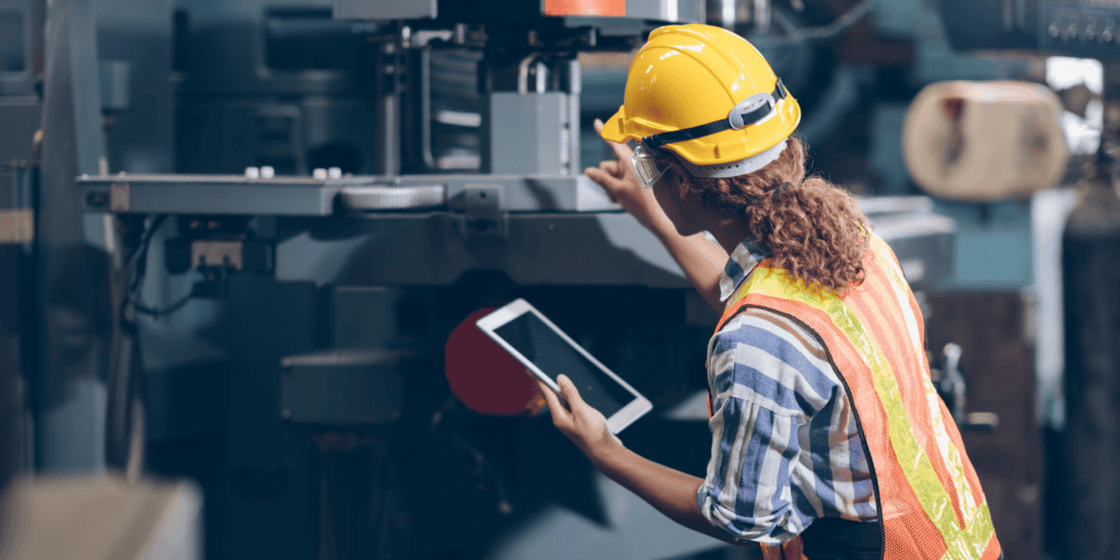 A person wearing a yellow hard hat, safety vest, and striped shirt is holding a tablet and inspecting machinery in an industrial or factory setting. The background is filled with industrial equipment and machinery.