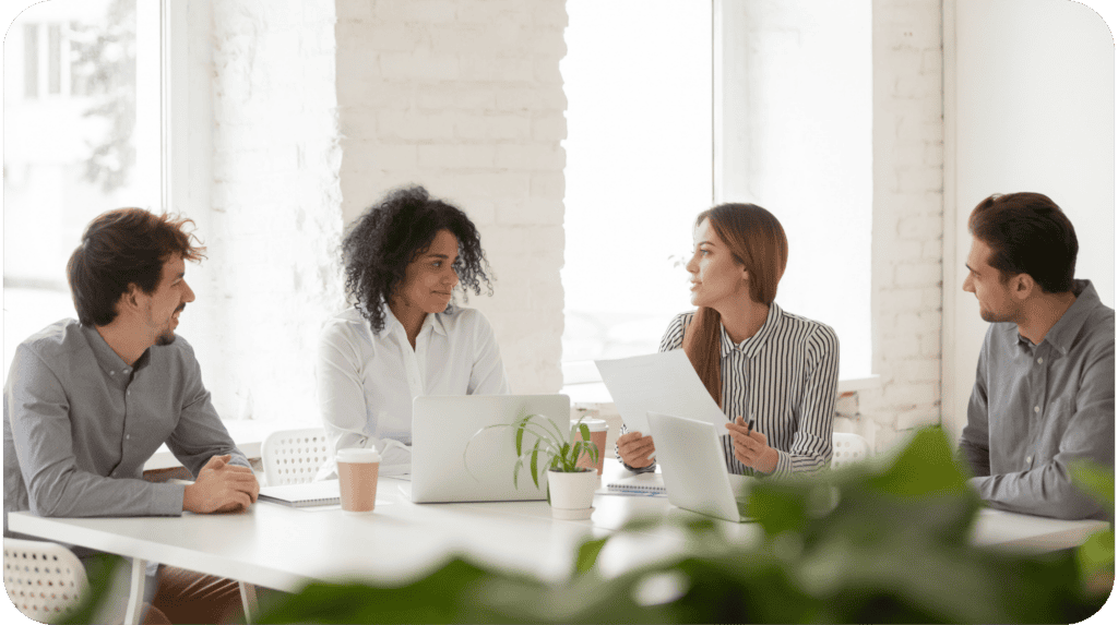 Four people sitting around a table and engaged in a meeting. Two laptops, a coffee cup, and a plant are on the table. The woman on the right is holding a piece of paper, while the others listen attentively. The setting appears to be a bright, modern office.