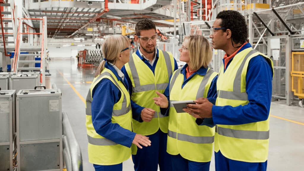 Four workers in high-visibility vests and safety glasses stand in an industrial facility, engaged in discussion. They appear to be collaborating on a task, with one holding a tablet. The background shows machinery and industrial equipment.