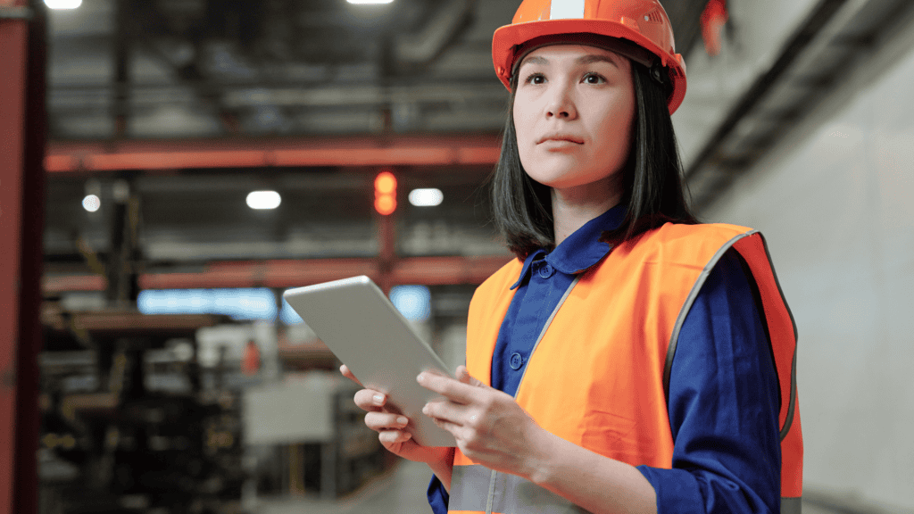 A woman wearing an orange hard hat and safety vest stands in an industrial setting, holding a tablet. She looks upward with a serious expression. Stacks of materials and machinery are visible in the blurred background.