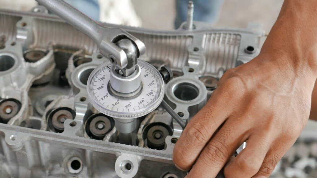 A person's hand adjusts a mechanical gauge positioned on an automotive engine part, using a ratchet wrench. The gauge displays angular measurements, and various engine components are visible around the gauge.