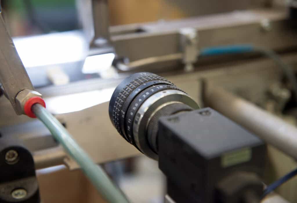 Close-up view of industrial machinery featuring a metal component and a black cylindrical object with a lens, possibly a camera or sensor, surrounded by various tubes and mechanical parts.