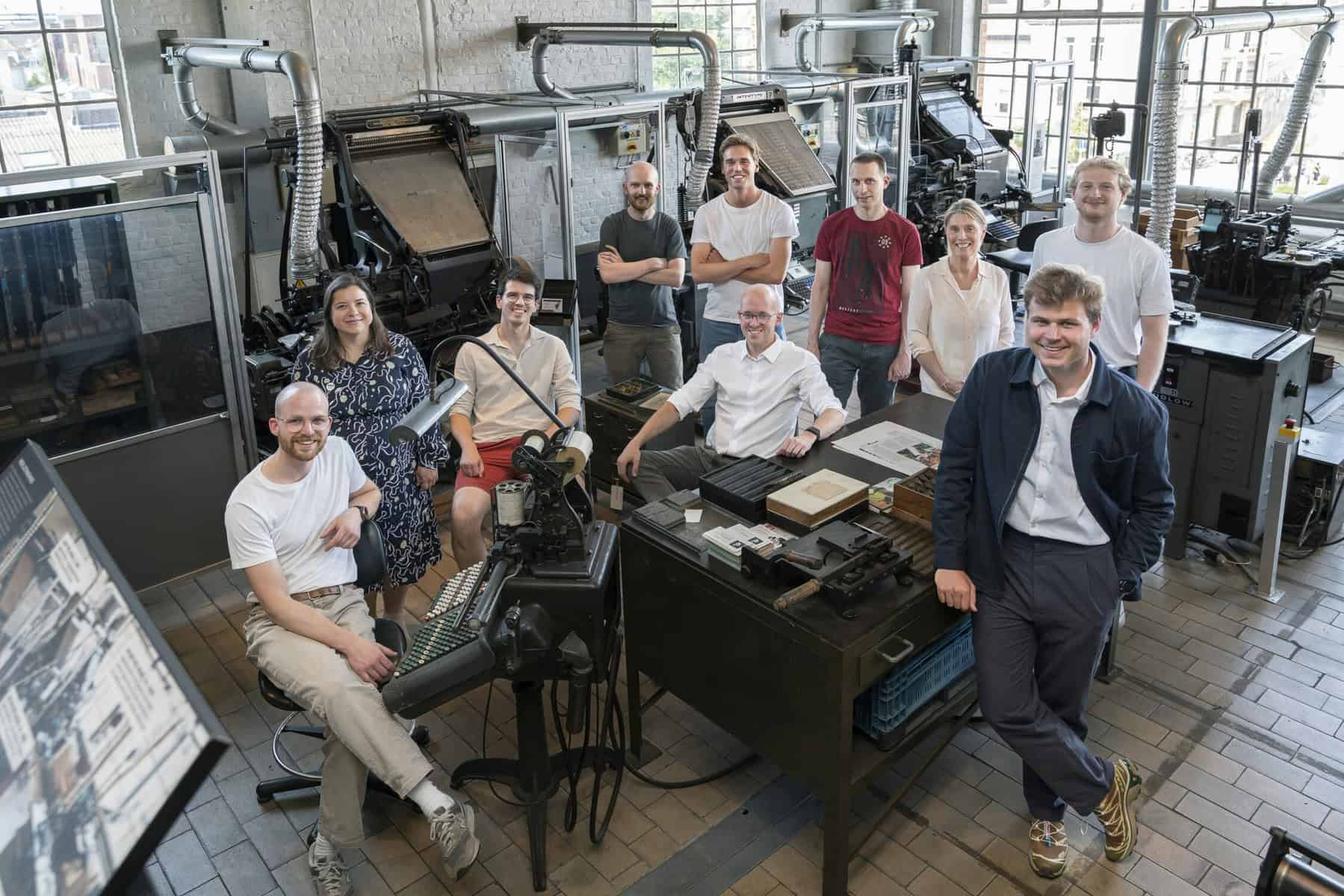 A group of ten people, composed of eight men and two women, stand and sit in a room filled with vintage printing machinery. They appear relaxed and are either smiling or looking at the camera. The setting has a workshop-like atmosphere with various machines and equipment.