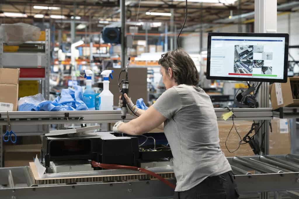 A person with short hair is operating machinery in a factory or industrial setting. Various tools, cleaning supplies, and boxes are on the workstation. A computer monitor near the person displays technical information or images. The background shows shelves and equipment.