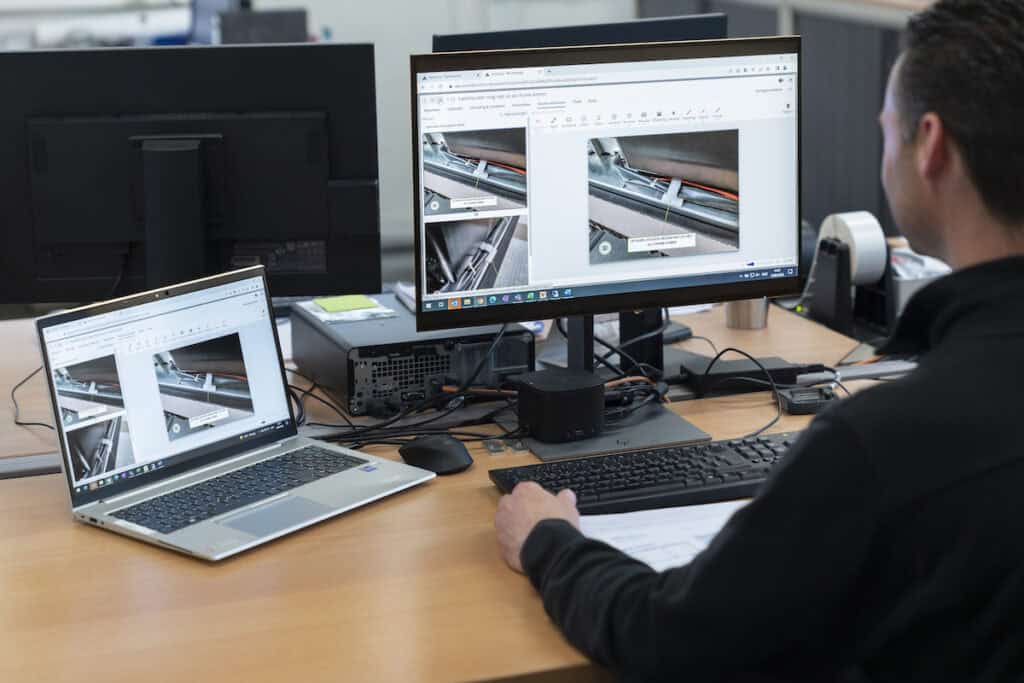 A person wearing a dark top is sitting at a desk with a laptop and a monitor. Both screens display technical images and documents. The desk has various office supplies, including a keyboard, mouse, and docking station. The background includes additional monitors.